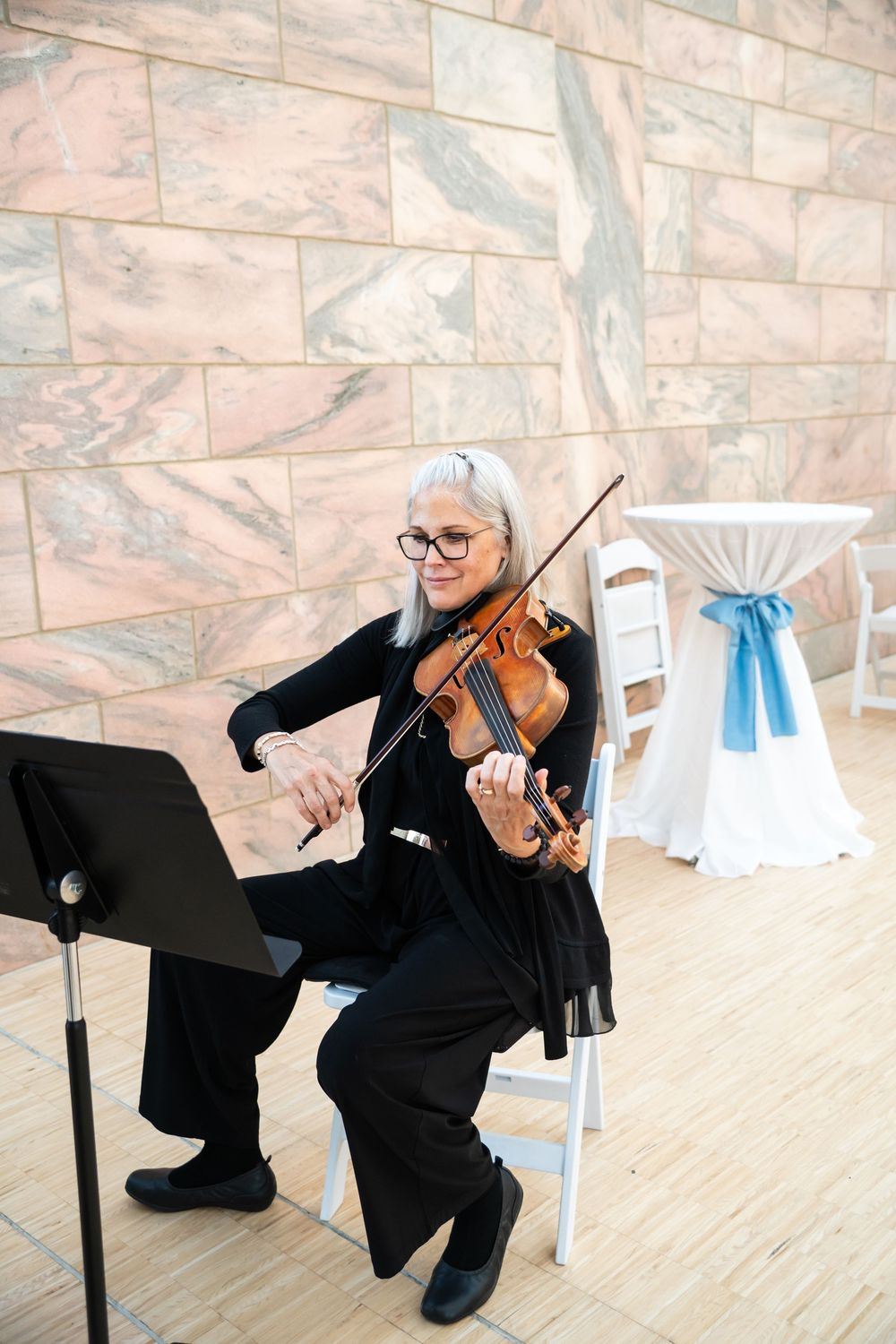 Elderly woman with gray hair playing violin while seated, wearing black outfit and glasses, performing at an elegant event.