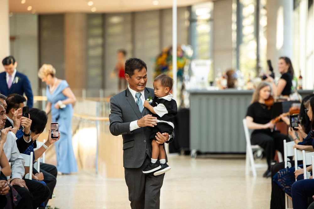 Groom in gray suit carries baby down wedding aisle while guests photograph the touching moment.