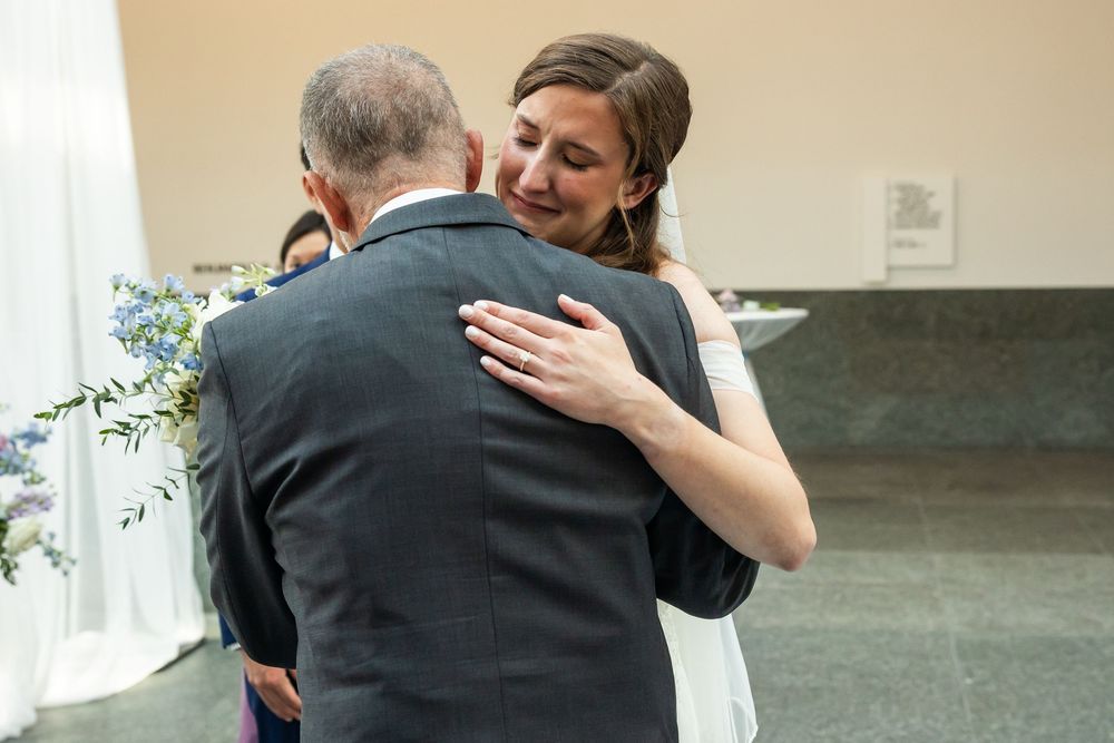 Bride in white gown emotionally hugging an older man in gray suit during wedding ceremony indoors.