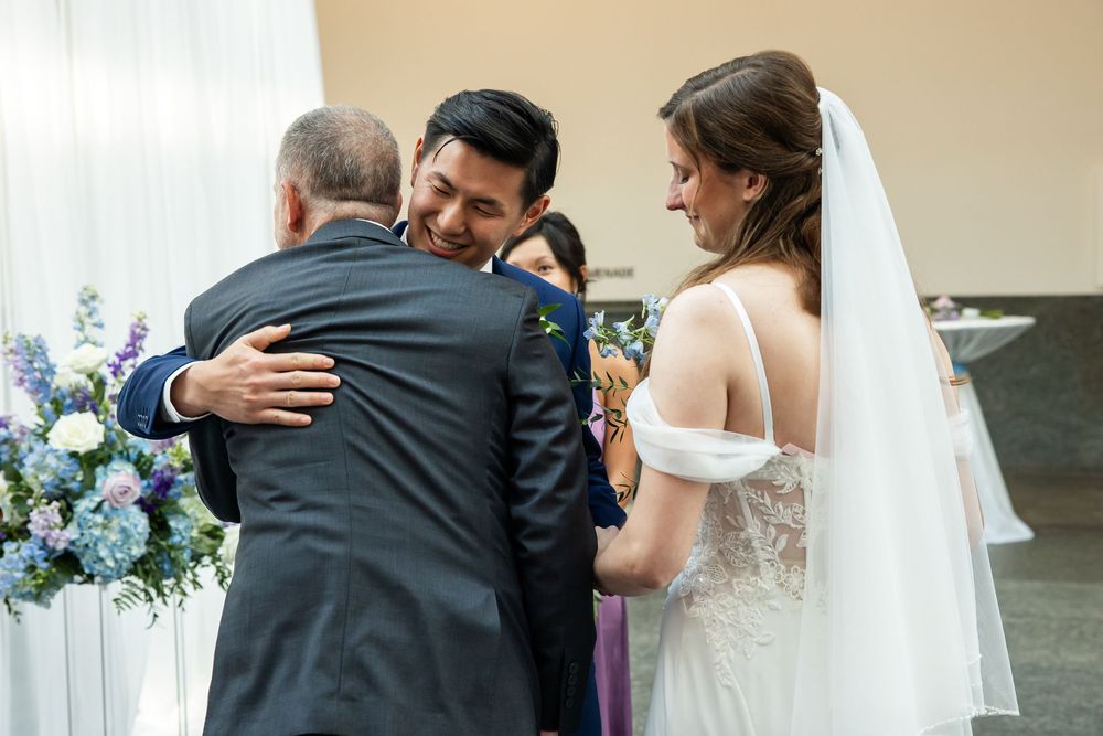 Groom hugging a guest at wedding ceremony while smiling bride in white gown and veil watches nearby.