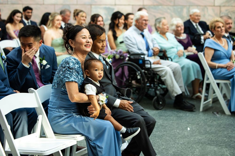 A woman in blue holds a young boy at an outdoor wedding ceremony with guests seated in white chairs.