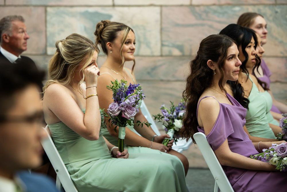 Bridesmaids in mint and purple dresses holding floral bouquets, one wiping tears during an emotional wedding ceremony.