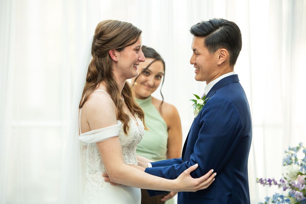 Bride and groom smiling at each other during wedding ceremony with officiant in background.