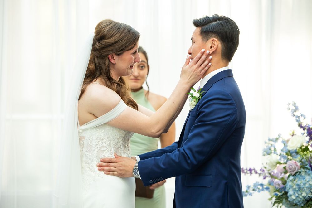Bride tenderly touches groom's face during wedding ceremony while officiant stands behind them.