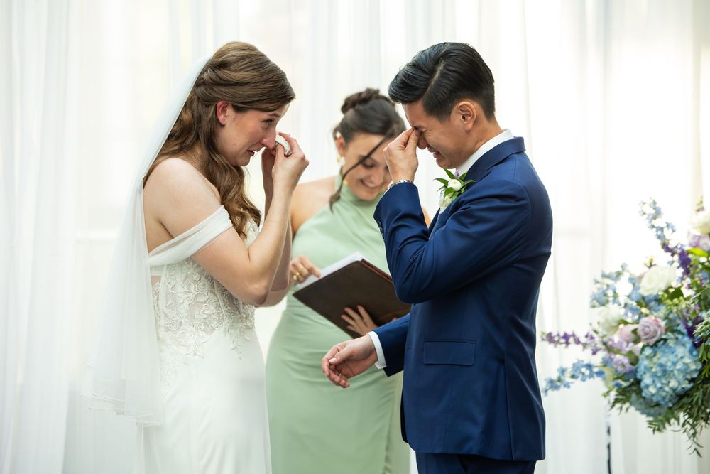 Emotional bride and groom crying during heartfelt wedding vows ceremony with officiant in elegant white venue.