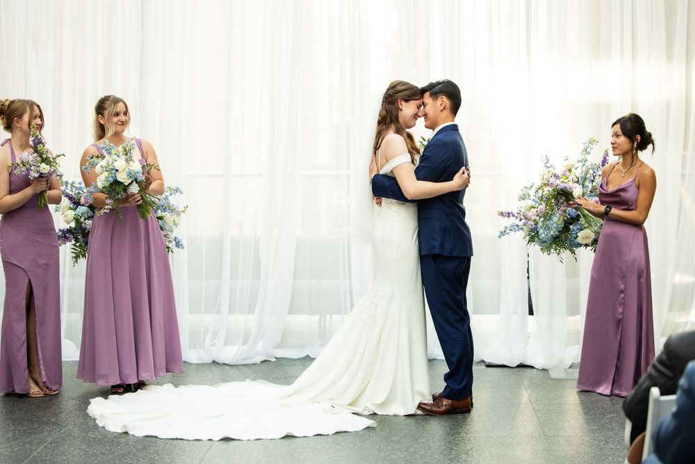 Bride and groom share first kiss at indoor wedding ceremony with bridesmaids in purple gowns holding floral bouquets.
