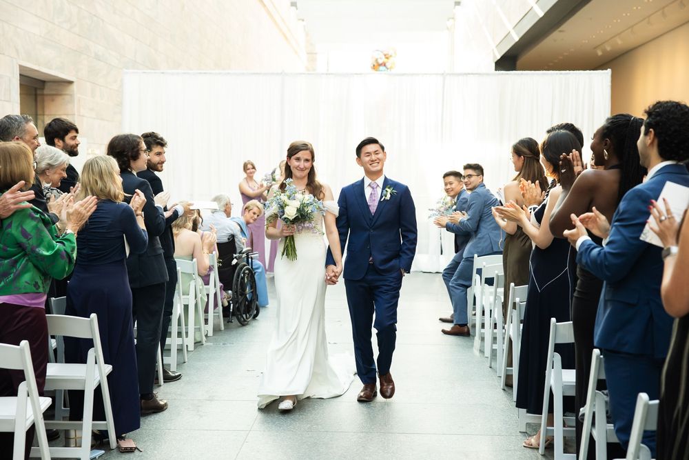 Newlywed couple walks down wedding aisle hand-in-hand as guests applaud in elegant white ceremony venue.