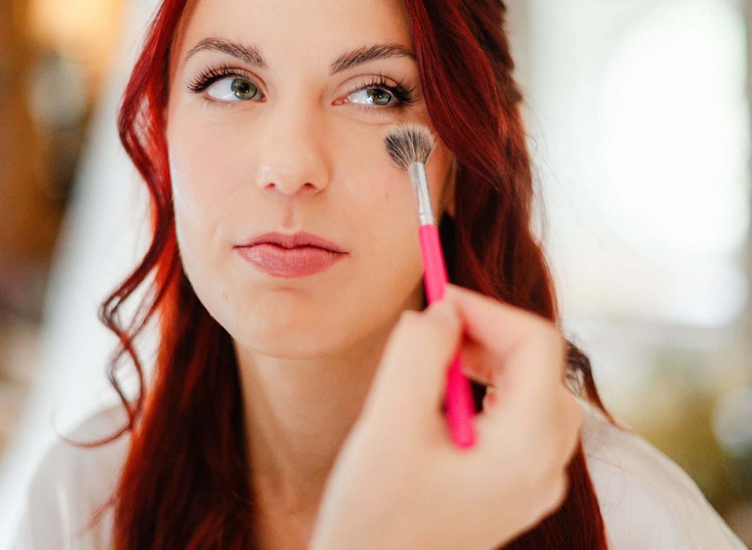 Red-haired woman getting eye makeup applied with a pink brush by a professional makeup artist.