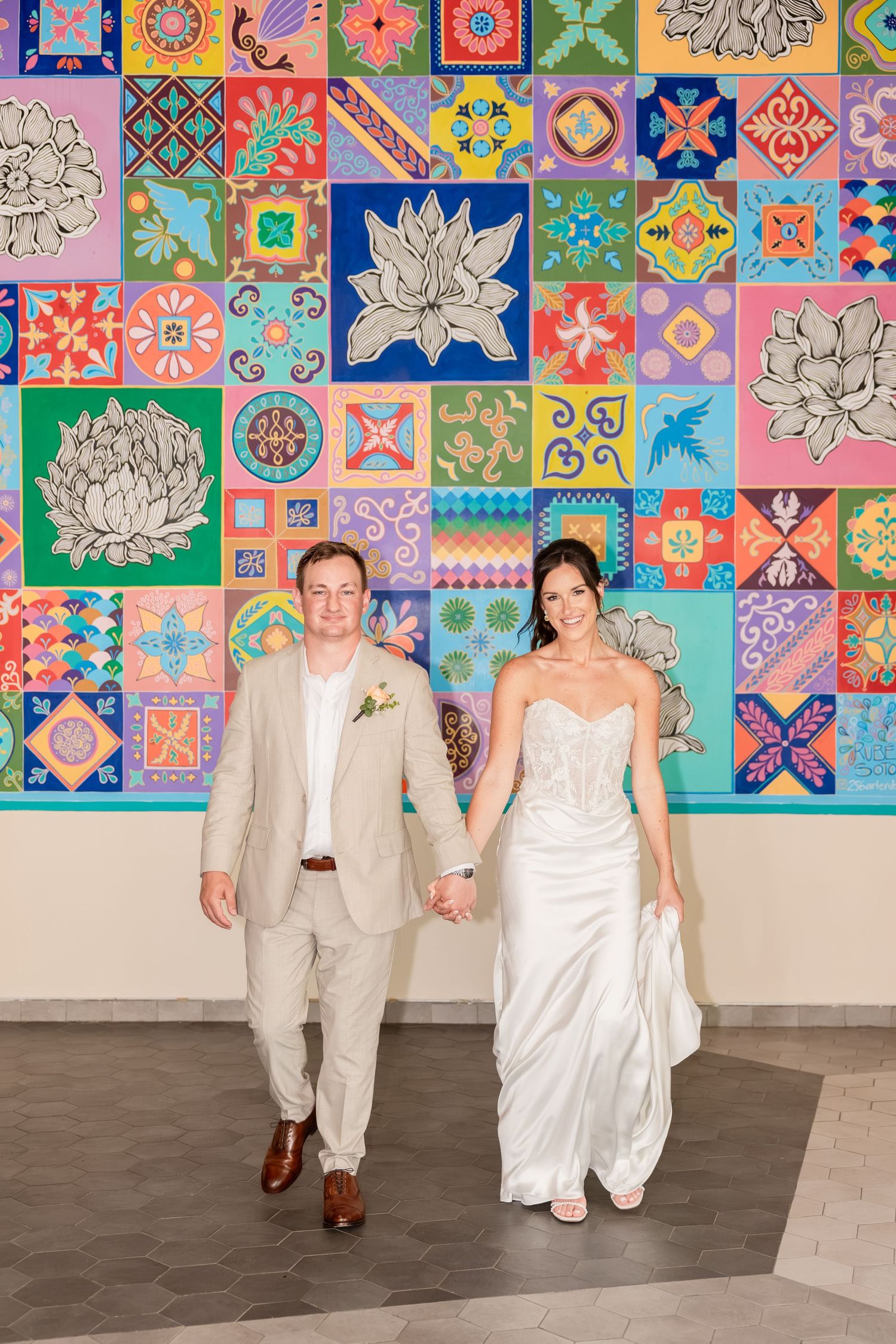 Bride and groom hold hands walking in front of colorful mosaic tile wall mural at wedding venue.