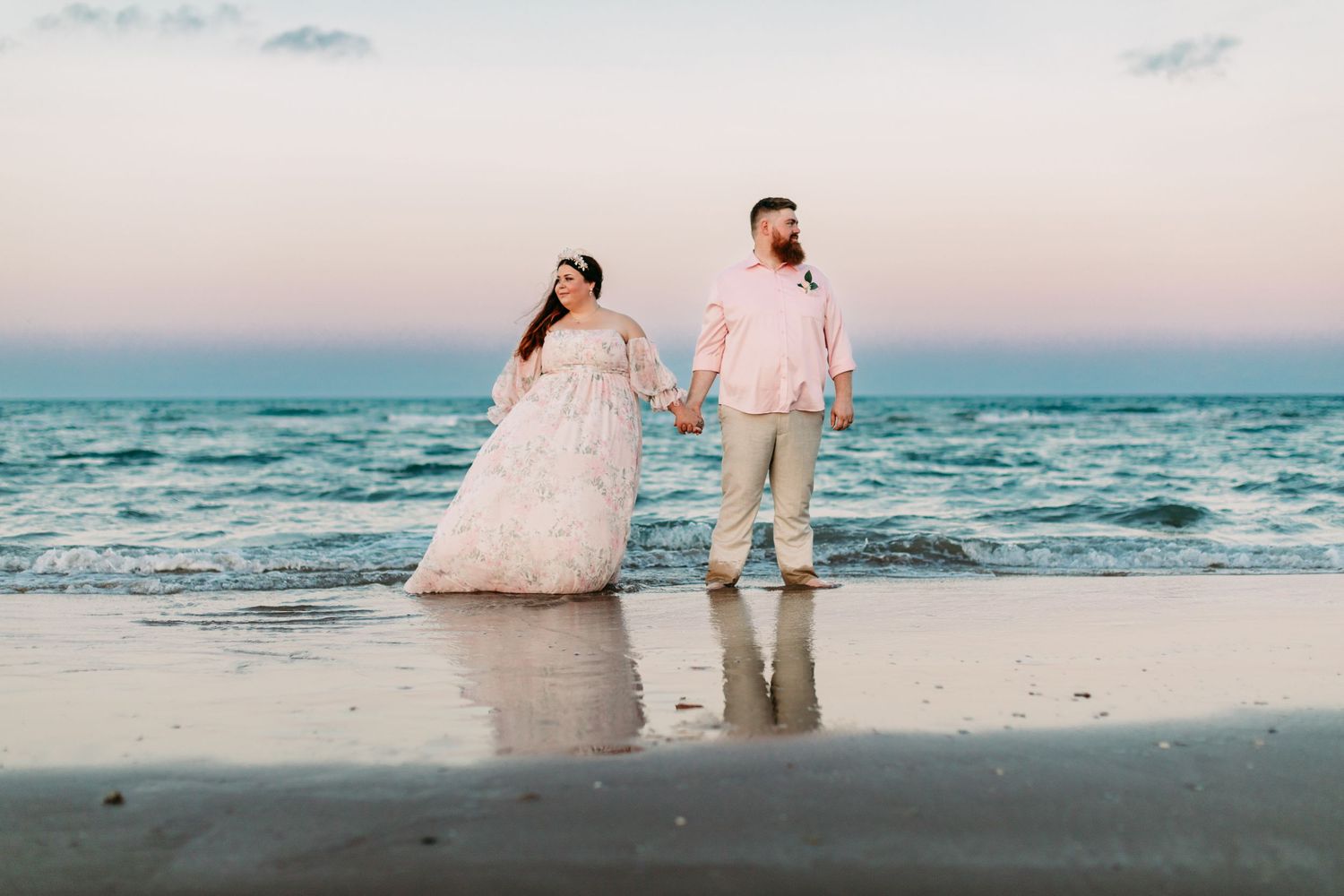 Bride and groom hold hands on a serene beach at sunset, waves reflecting pastel pink and blue sky.