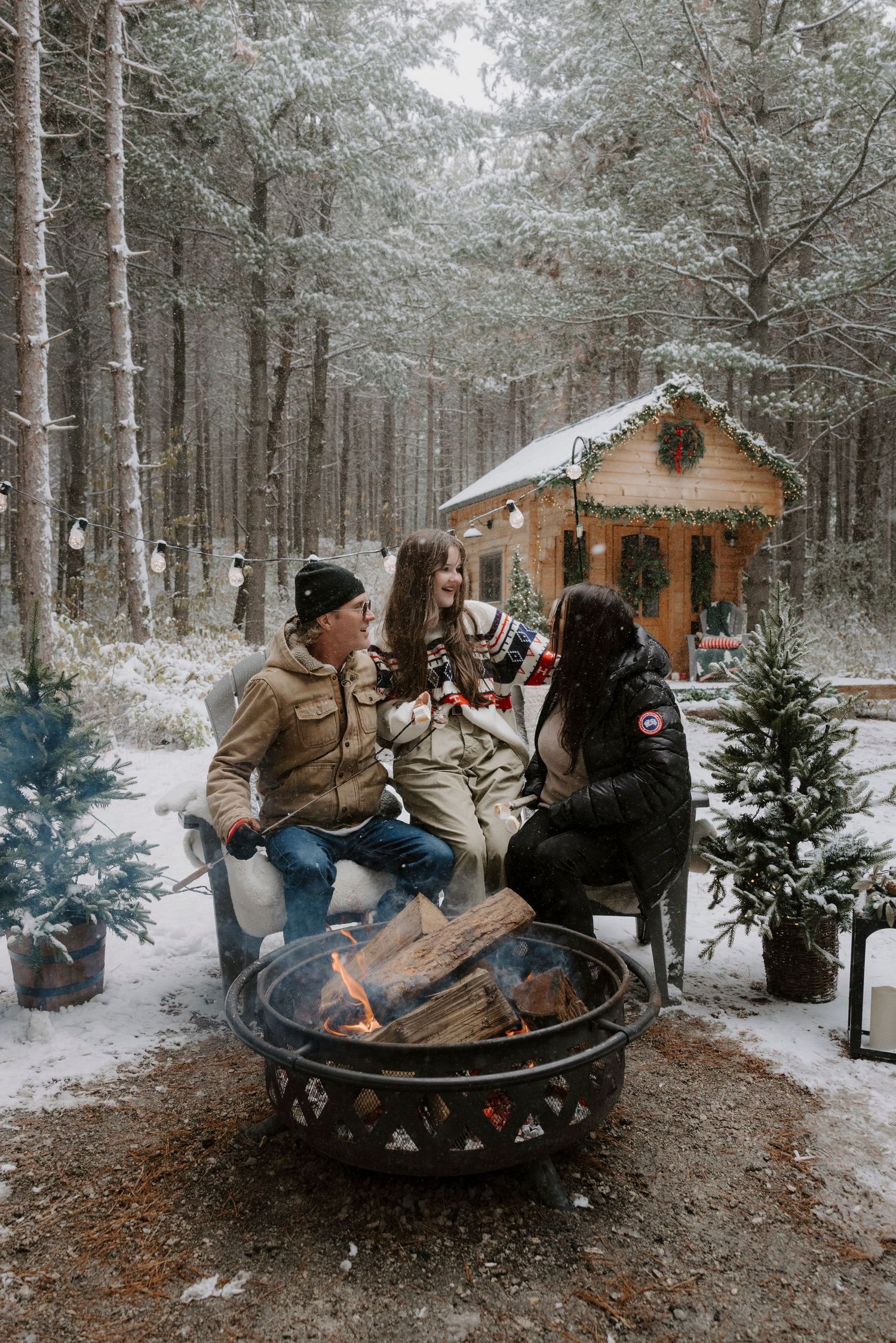 Three friends gather around a fire pit outside a snow-covered log cabin in a winter forest holiday setting.