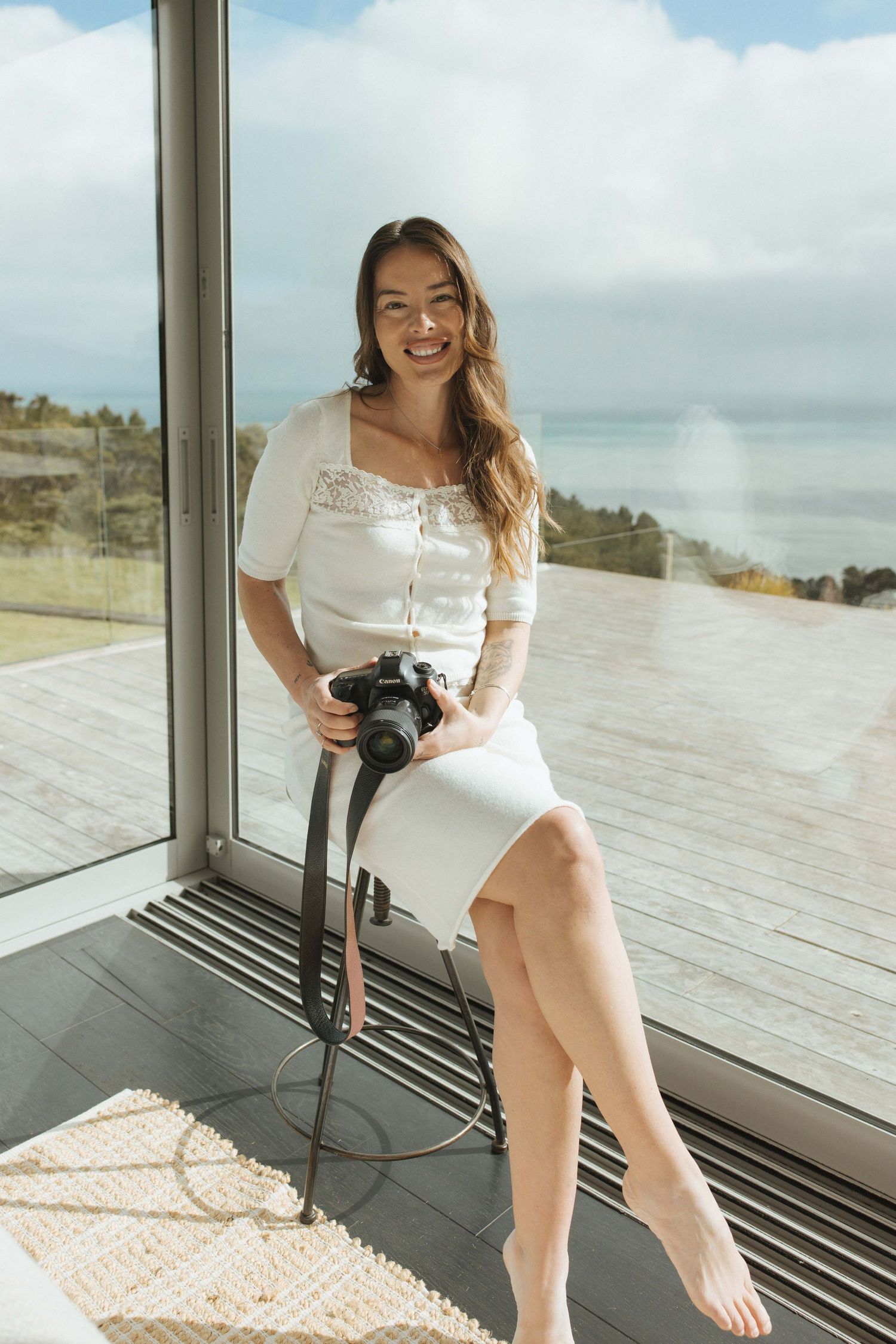 Smiling woman in white dress holding a DSLR camera, sitting by large glass windows with ocean view.