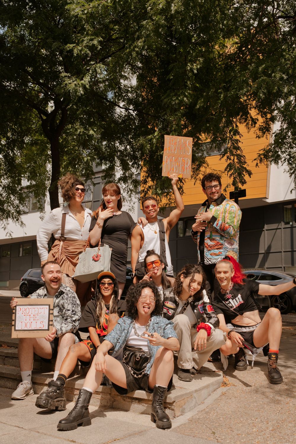 Group of young diverse friends posing outdoors holding protest signs, smiling and having fun near trees and a building.