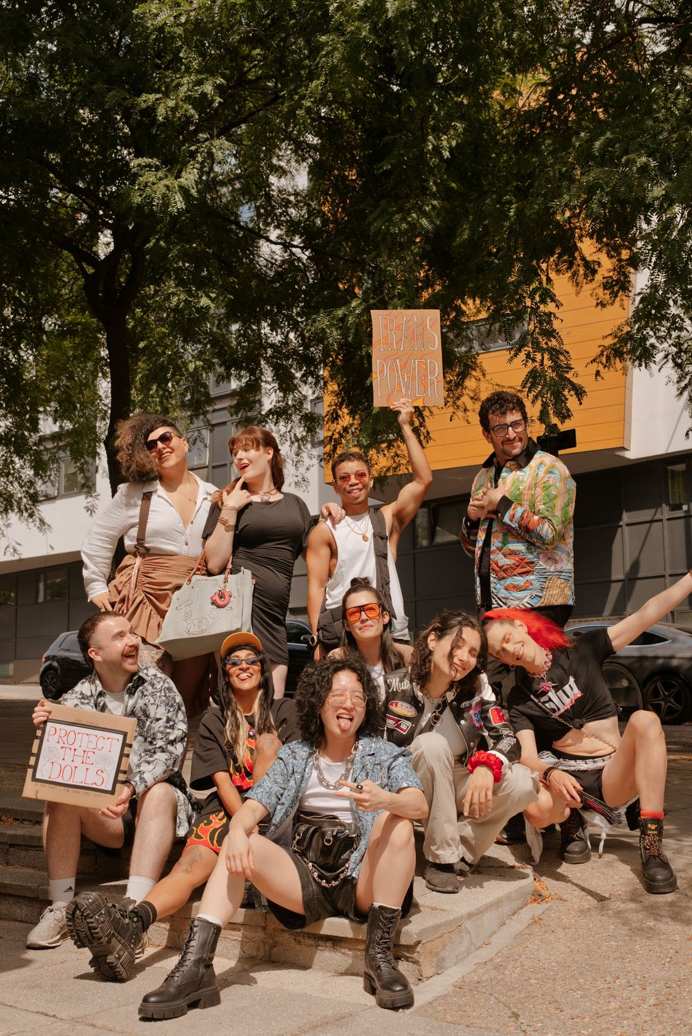 Diverse group of young people posing together outdoors on steps, holding signs, wearing colorful outfits on a sunny day.