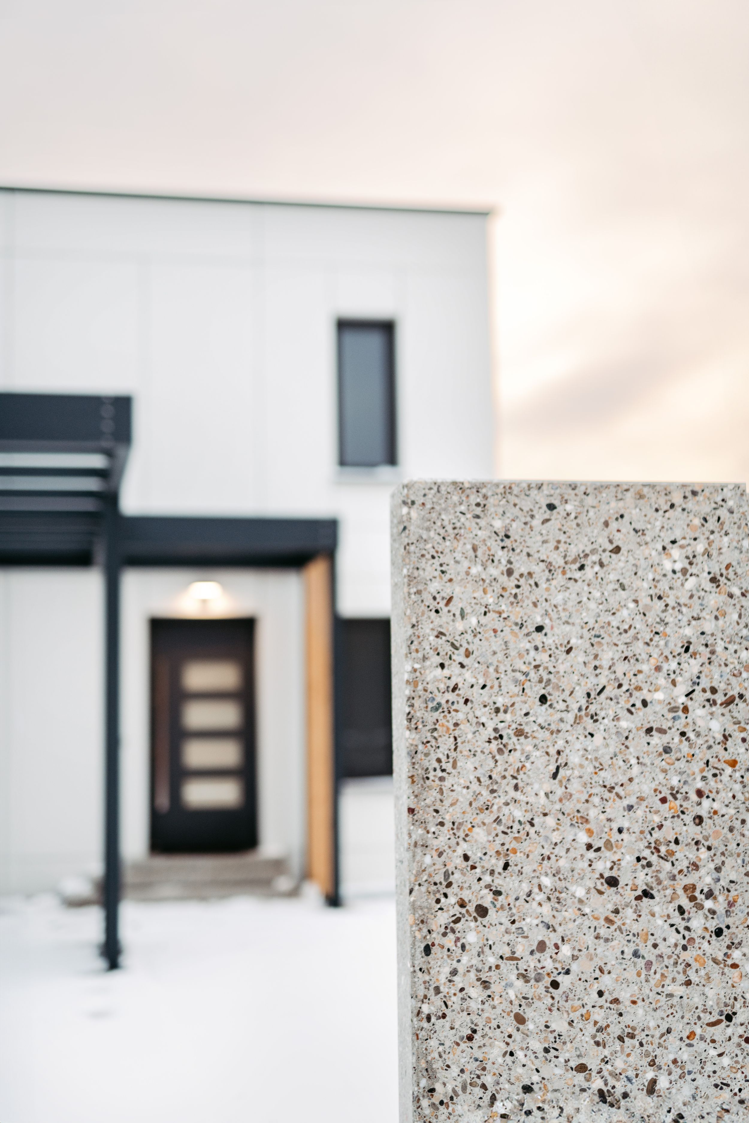 Modern white house with black accents and wooden front door, viewed behind a textured granite pillar in winter.