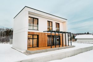 Modern two-story white house with wood accents and glass canopy, surrounded by snow in winter landscape.