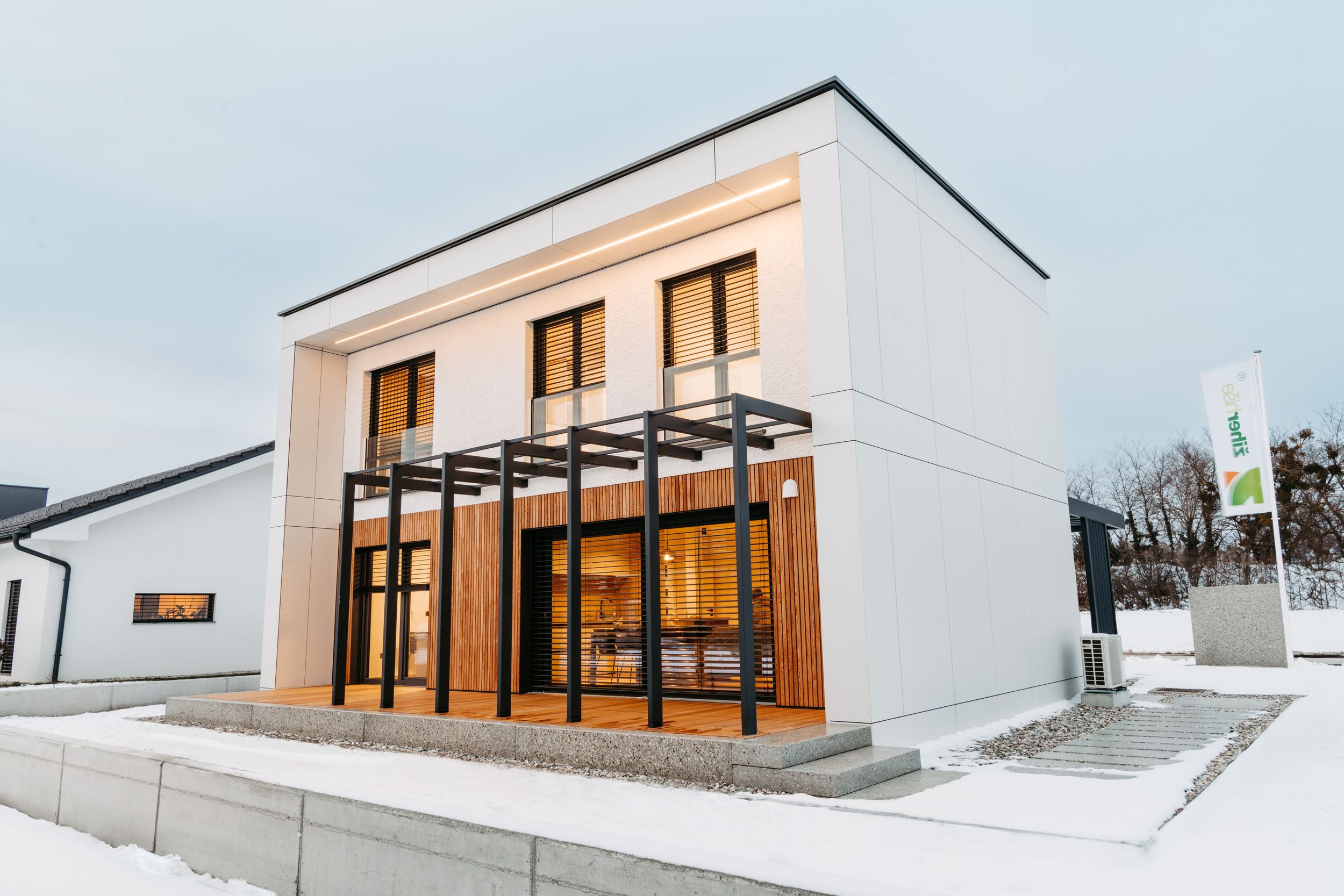Modern white two-story house with wood-accented glass entrance and flat roof covered in snow during winter.