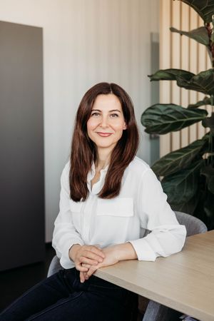 A smiling brunette woman in a white blouse sits at a wooden table in a modern office with a green plant.