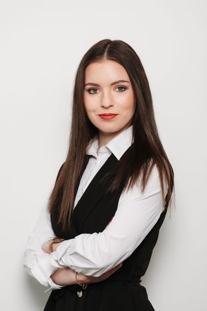 Young professional woman with long brown hair in black vest and white shirt, arms crossed, posing against white background.