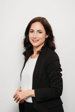 Professional woman in black blazer and white top smiling confidently against a light gray background.