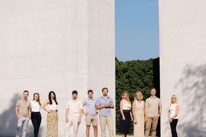 Group of nine professionals posing between white stone columns outdoors on a sunny day.