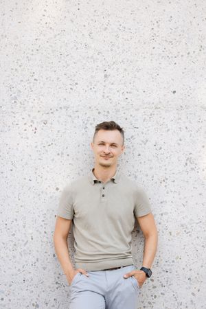 Young man in beige polo shirt leaning against a textured concrete wall with hands in pockets, smiling confidently.