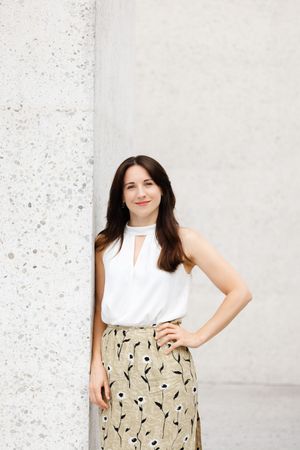 Woman in white sleeveless top and floral skirt leaning against a concrete column, smiling confidently.
