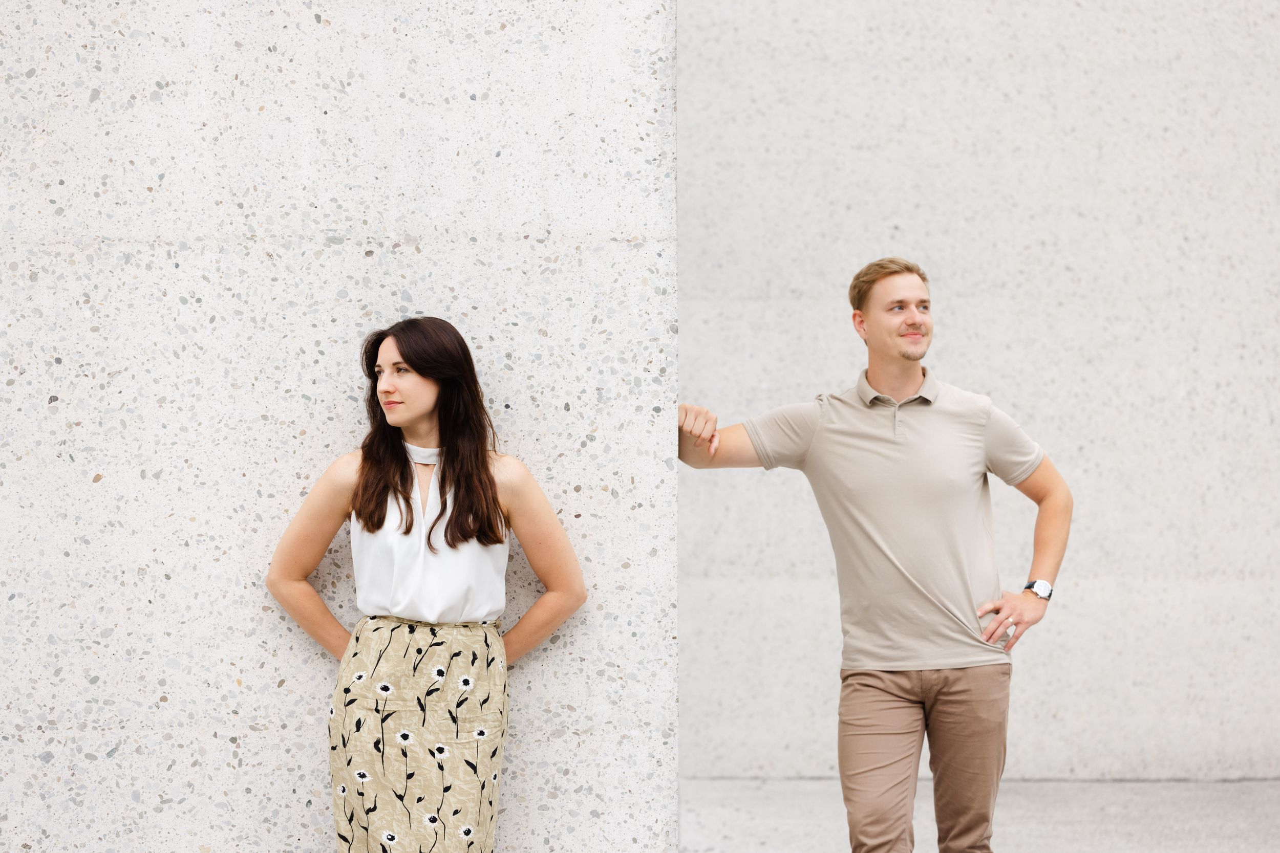 A woman in a floral skirt and a man in a beige polo pose separately against a light concrete wall outdoors.