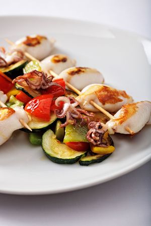 Grilled squid and vegetable skewers with zucchini, red peppers, and baby octopus on a white plate.