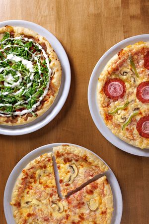 Three pizzas on white plates on a wooden table, topped with arugula, salami, mushrooms, and melted cheese.