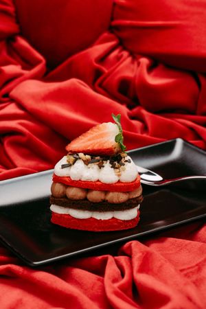 Elegant red velvet layered dessert topped with fresh strawberry and whipped cream on black plate against red silk fabric.