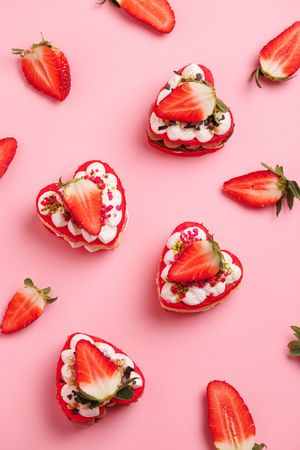 Heart-shaped strawberry shortcake desserts topped with cream and fresh strawberries on a pink background.