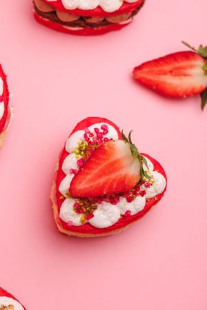 Heart-shaped strawberry tart with whipped cream and pistachio crumbles on a pink background.