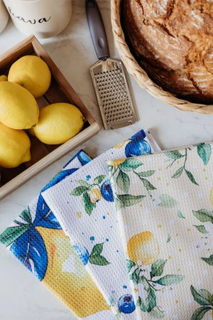 Lemon-printed kitchen towels on marble surface with fresh lemons in wooden tray, grater, and rustic bread in basket.