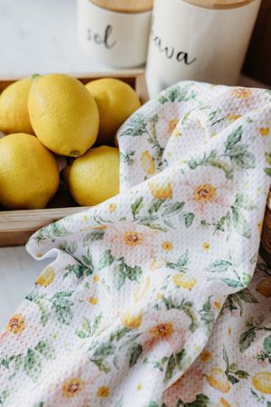 Floral kitchen towel with pink roses and lemons beside a wooden tray of fresh yellow lemons and white canisters.