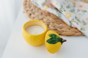 Yellow lemon-shaped ceramic candle holder with white wax, green leaf lid, wicker tray, and floral fabric in background.