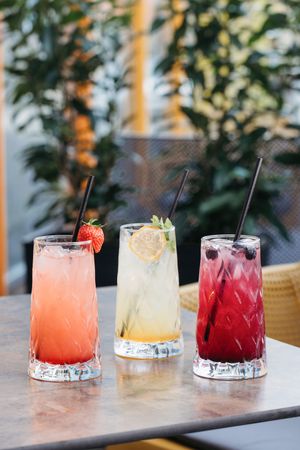 Three colorful cocktails with black straws on a table, featuring strawberry, lemon, and berry flavors, garnished with fresh fruit.
