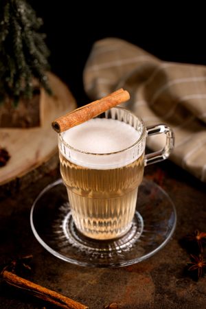 A warm spiced latte in a glass mug topped with foam and a cinnamon stick, surrounded by star anise on a dark surface.