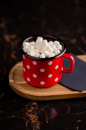 Red polka dot enamel mug filled with hot cocoa and mini marshmallows on a wooden board with dark background.