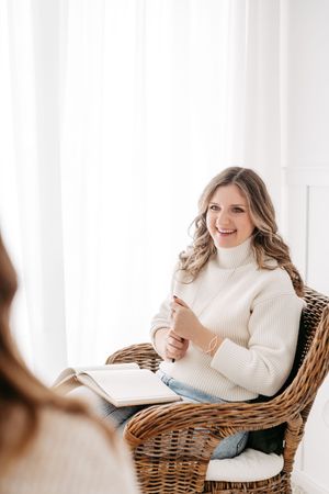 Smiling woman in white turtleneck sweater sitting in wicker chair holding pen and open notebook.