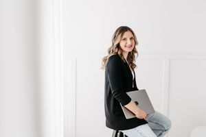 A smiling woman in a black cardigan and jeans holds a laptop while sitting in a bright white modern room.