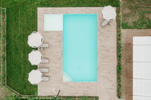 Aerial view of a rectangular turquoise swimming pool surrounded by wooden deck, white umbrellas, and lush green lawn.