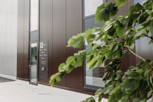 Modern building entrance with brown door panel, intercom system, and lush green foliage in foreground.