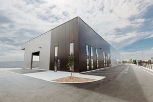 Modern industrial warehouse building with dark metal facade, large windows, and spacious parking lot under a partly cloudy sky.