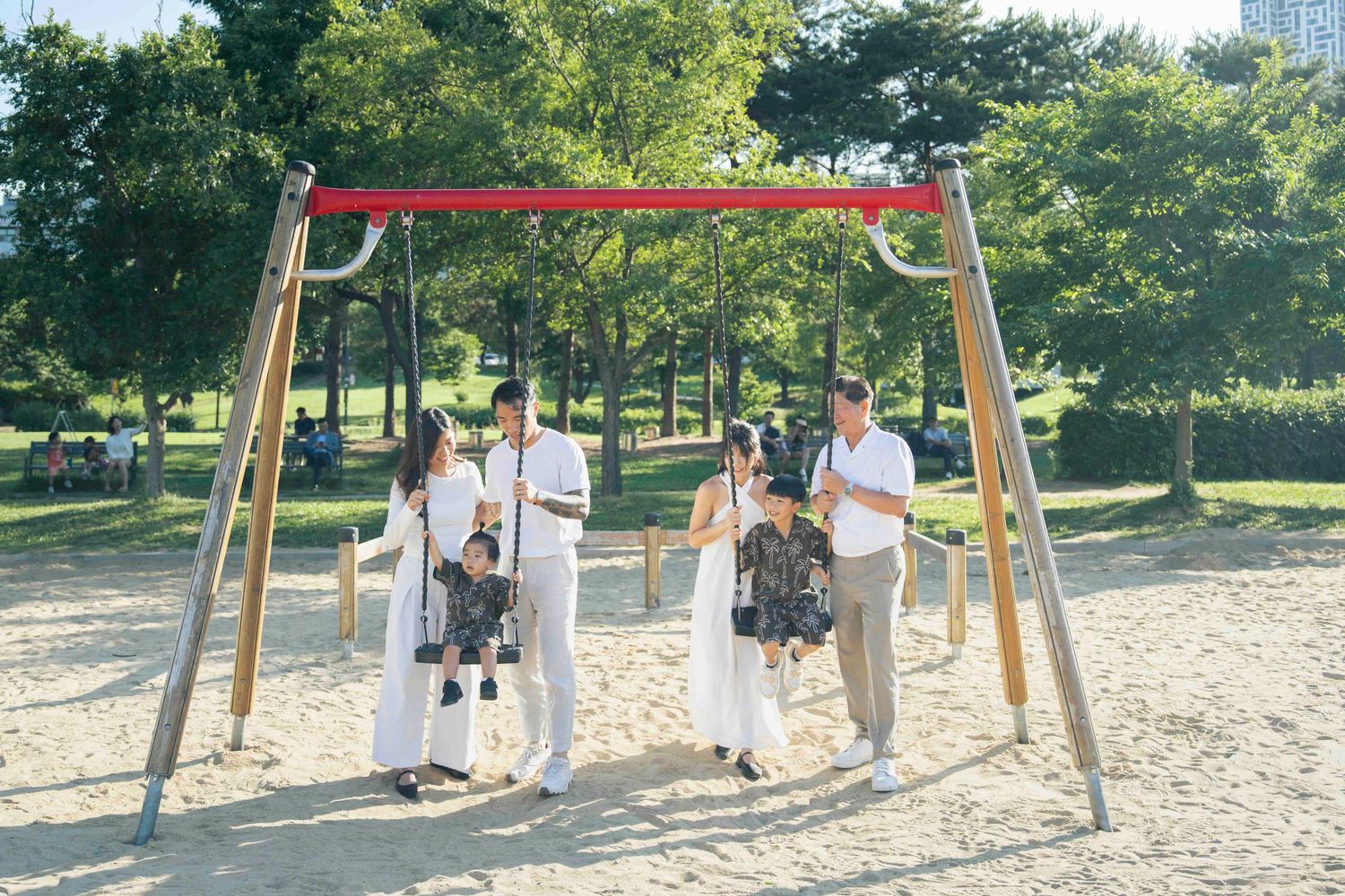 Two families with young children enjoy a sunny day at a park playground with wooden swing set on sandy ground.