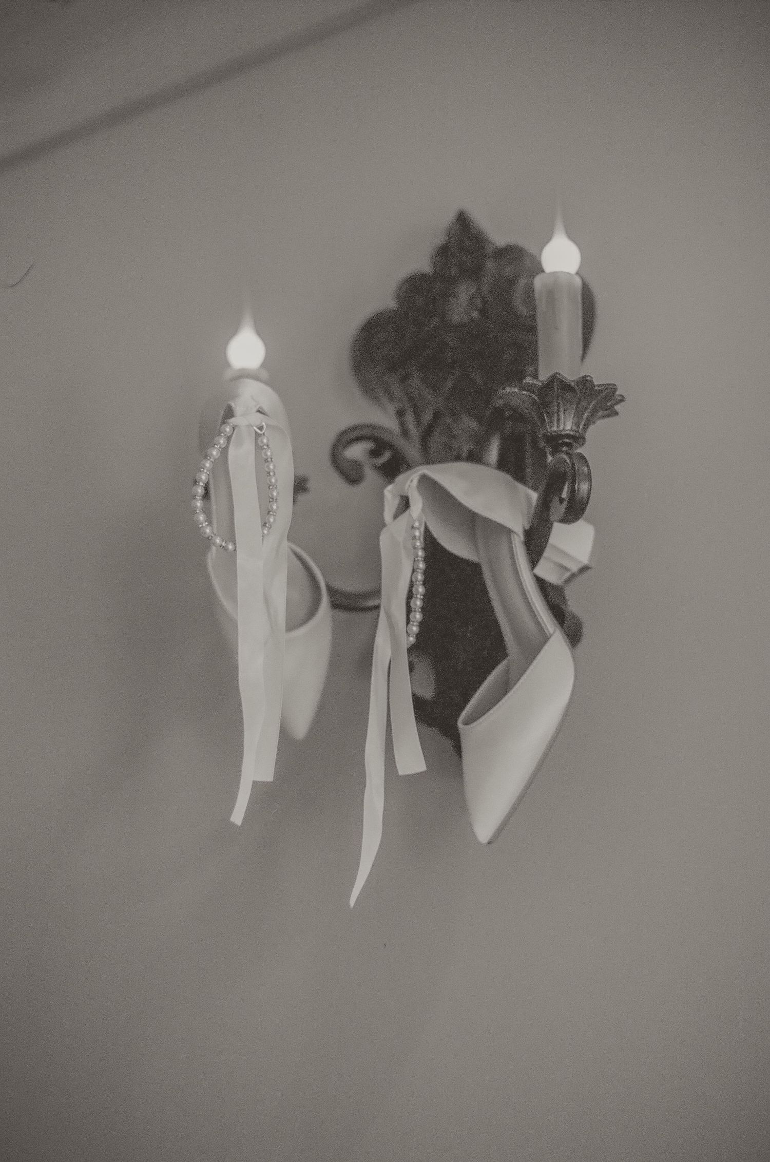 Elegant bridal heels with pearl details and ribbons hanging on ornate wall sconces in black and white photo.