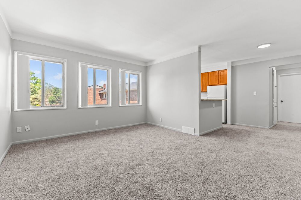 Bright empty living room with gray carpet, white walls, and a view into a kitchen with wood cabinets.