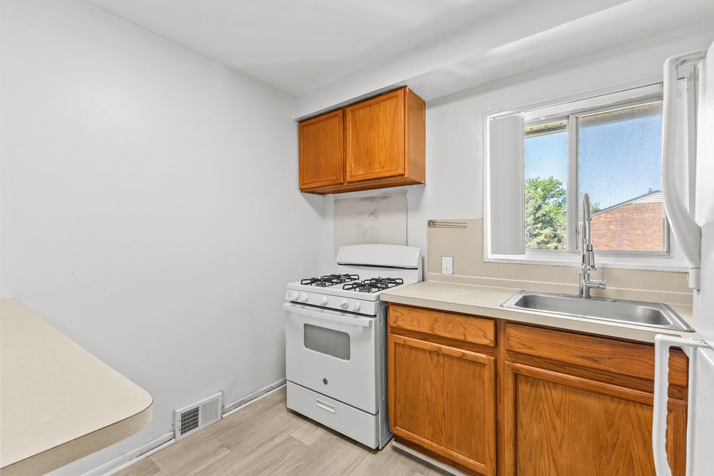 Small kitchen with oak cabinets, white gas range, and single basin sink near a bright window with light wood floors.