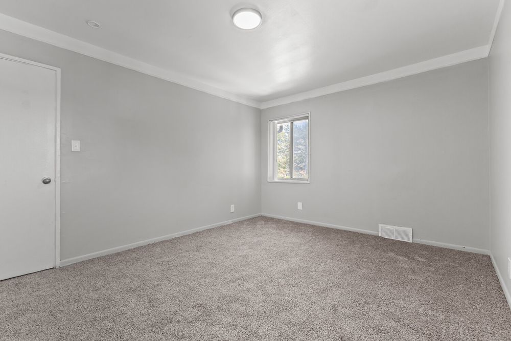 Empty carpeted bedroom with crown molding, neutral gray walls, small window, and a white door.