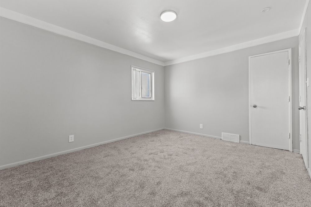 Bright empty bedroom featuring gray carpet, crown molding, white walls, and a single door.